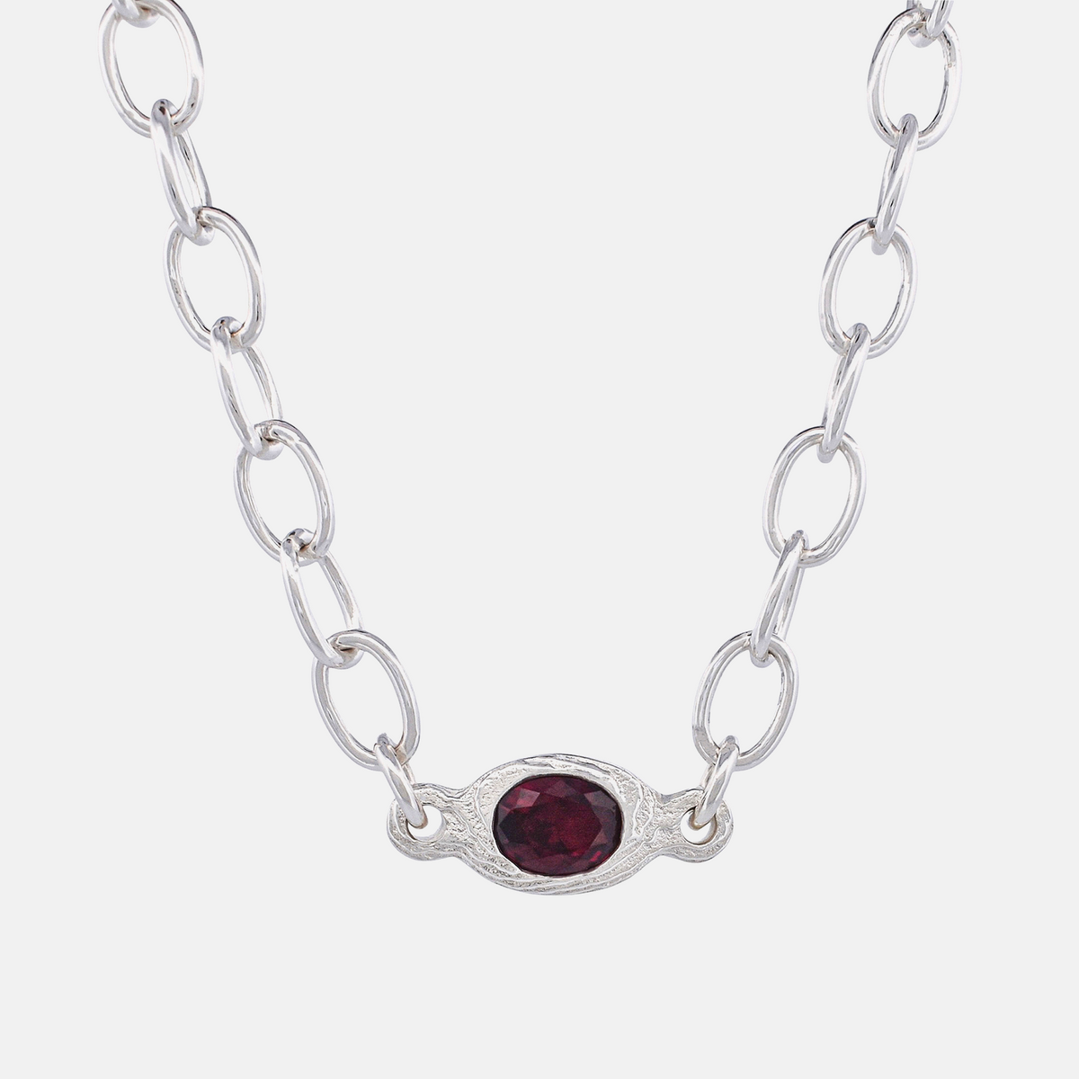 A silver link chain necklace featuring an oval silver pendant with a textured border and a faceted dark red gemstone in the center, set against a plain light gray background.