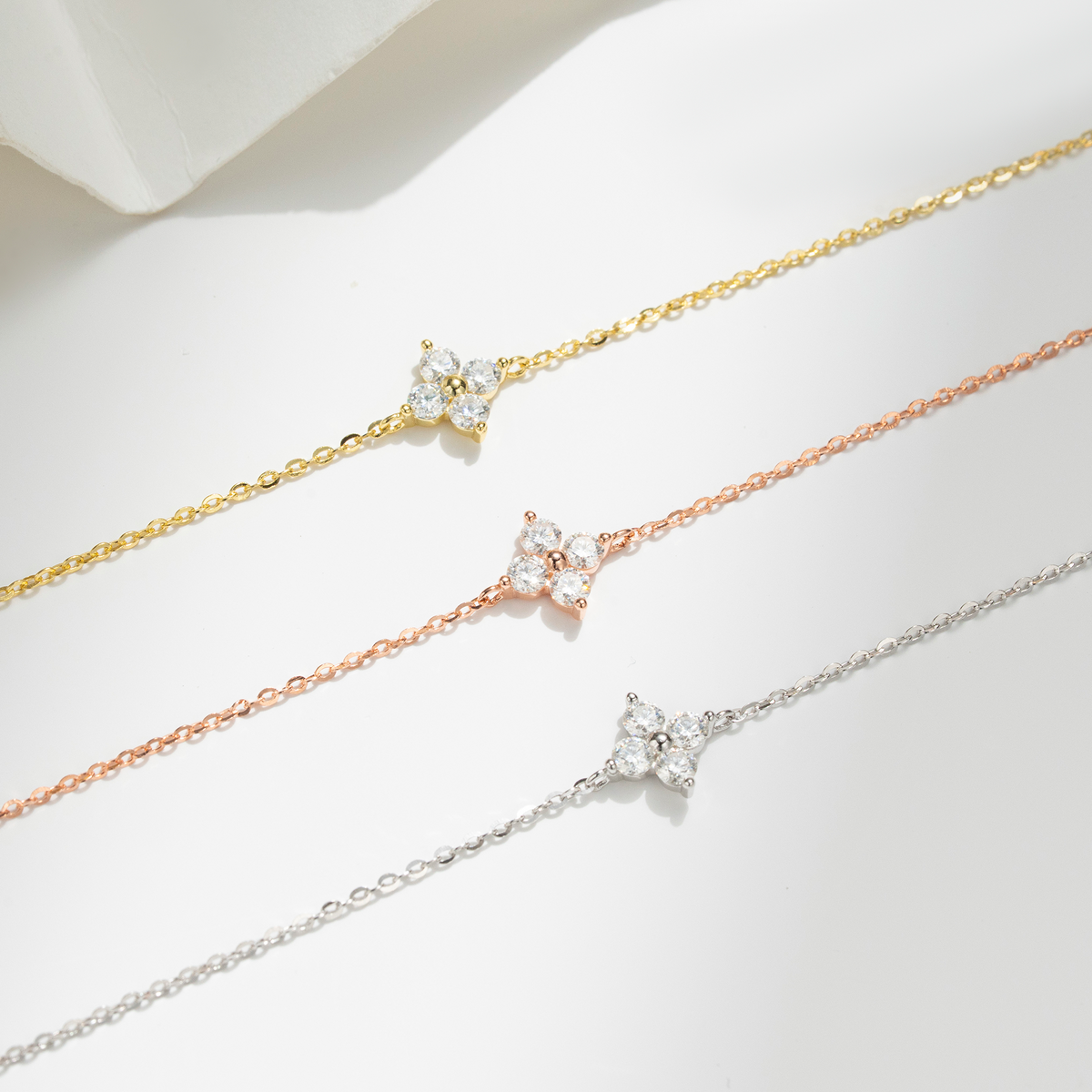 Three delicate chain bracelets in yellow gold, rose gold, and silver, each featuring a small flower-shaped cluster of clear cubic zirconia stones, arranged diagonally on a white surface.