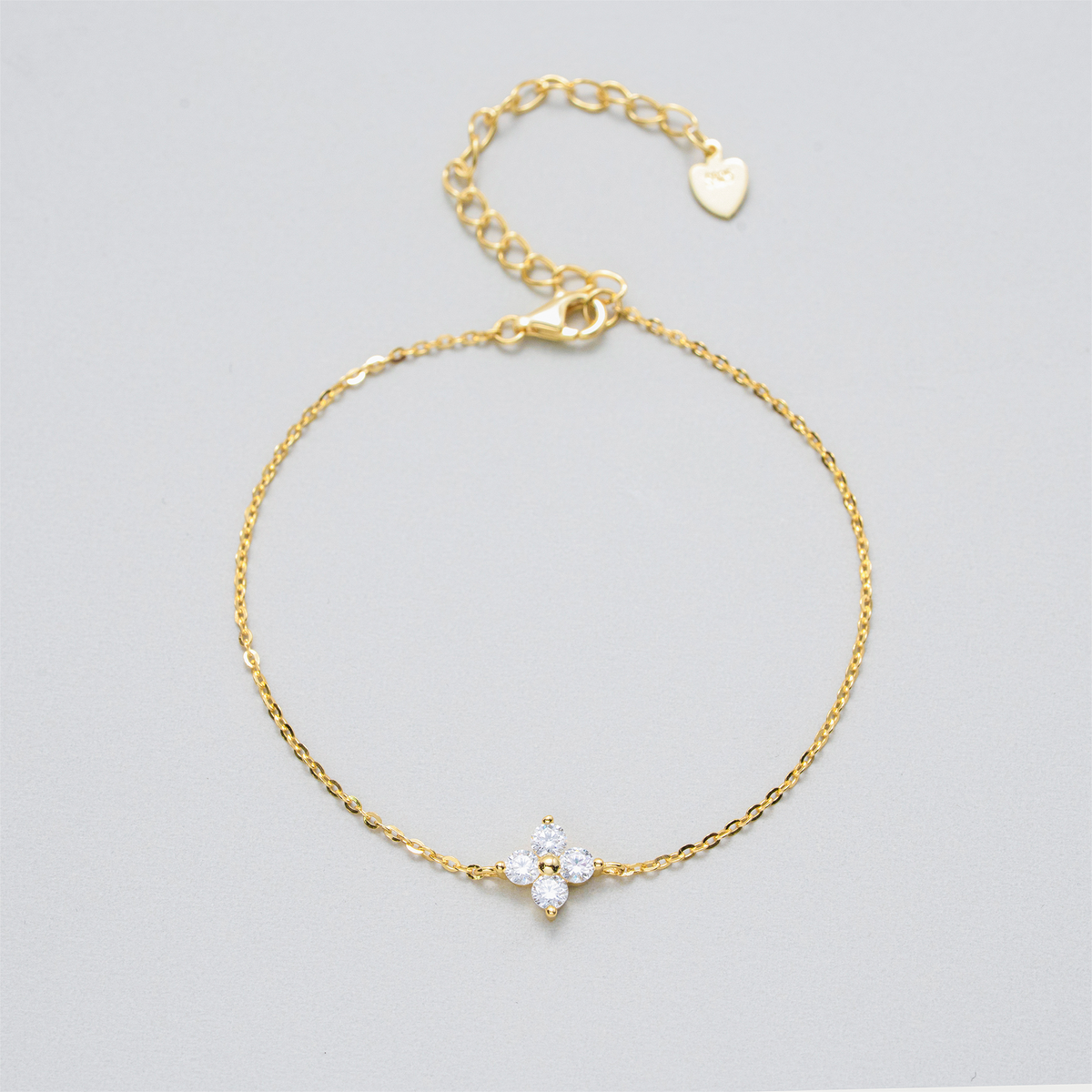 A delicate gold chain bracelet with a small flower-shaped charm set with clear stones, displayed on a light gray background.