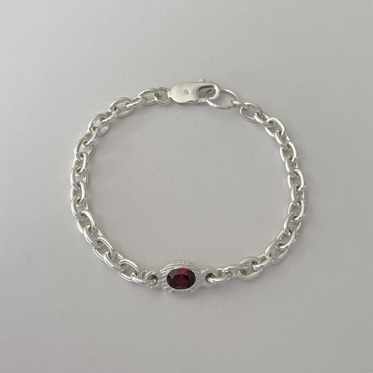 A silver chain bracelet with a small oval red gemstone centerpiece, displayed on a neutral light gray background.