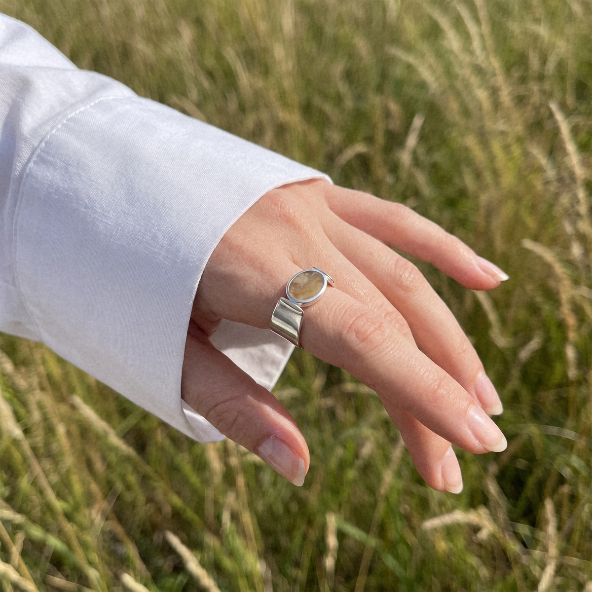 A silver ring with an oval natural stone worn on a person's ring finger, shown against a blurred background of tall green grass.