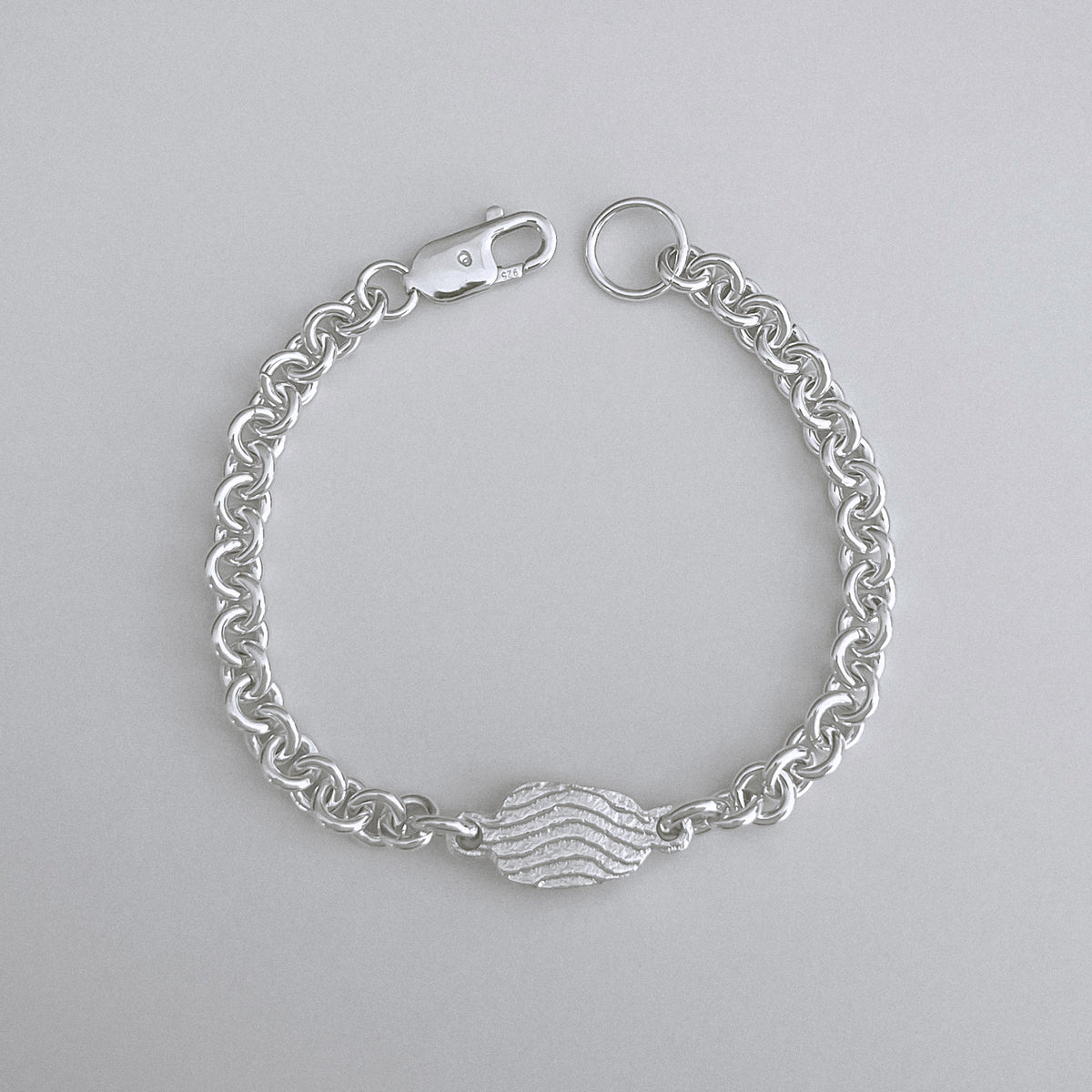 A silver chain bracelet with a central textured heart charm, displayed on a neutral light gray background.