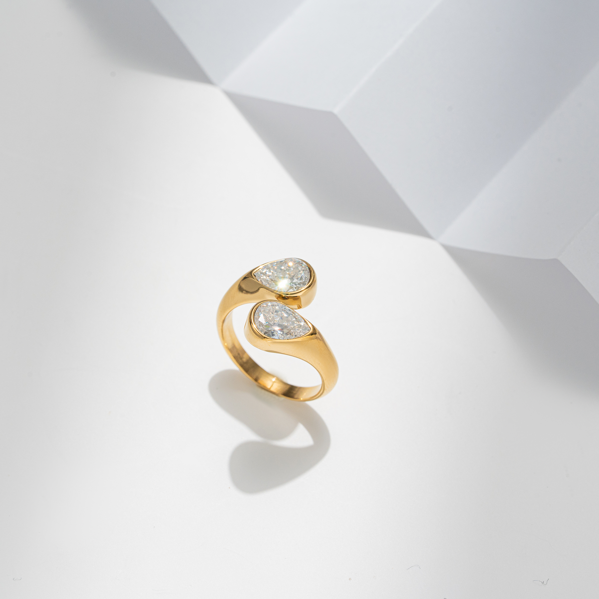 A gold ring with two pear-cut clear gemstones, photographed on a clean white geometric surface with soft shadows.