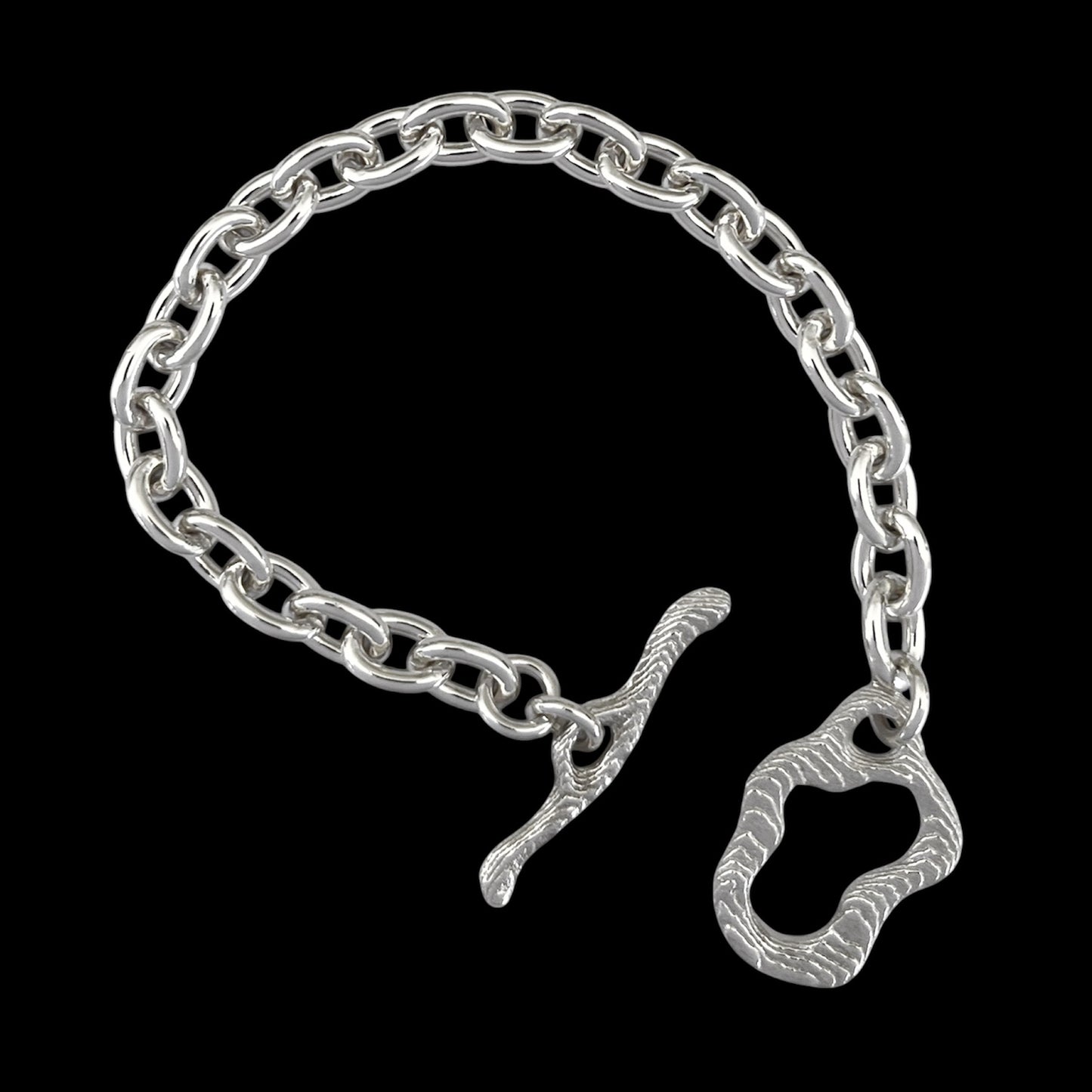 A silver chain bracelet with a unique textured toggle clasp and a matching decorative ring charm, displayed on a solid black background.