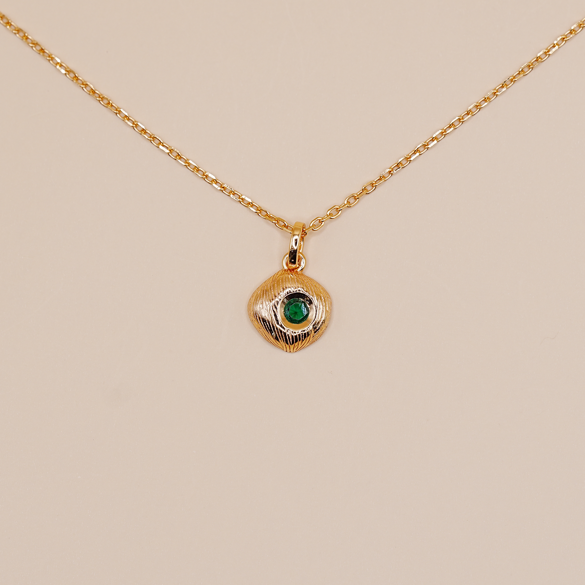 A delicate gold chain necklace featuring a small, textured gold pendant with a central green stone, displayed against a solid beige background.