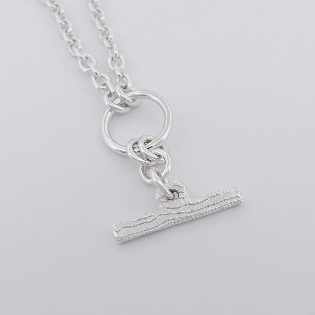 A silver pendant necklace featuring a textured horizontal bar charm suspended from a ring and chain, shown on a light gray background.