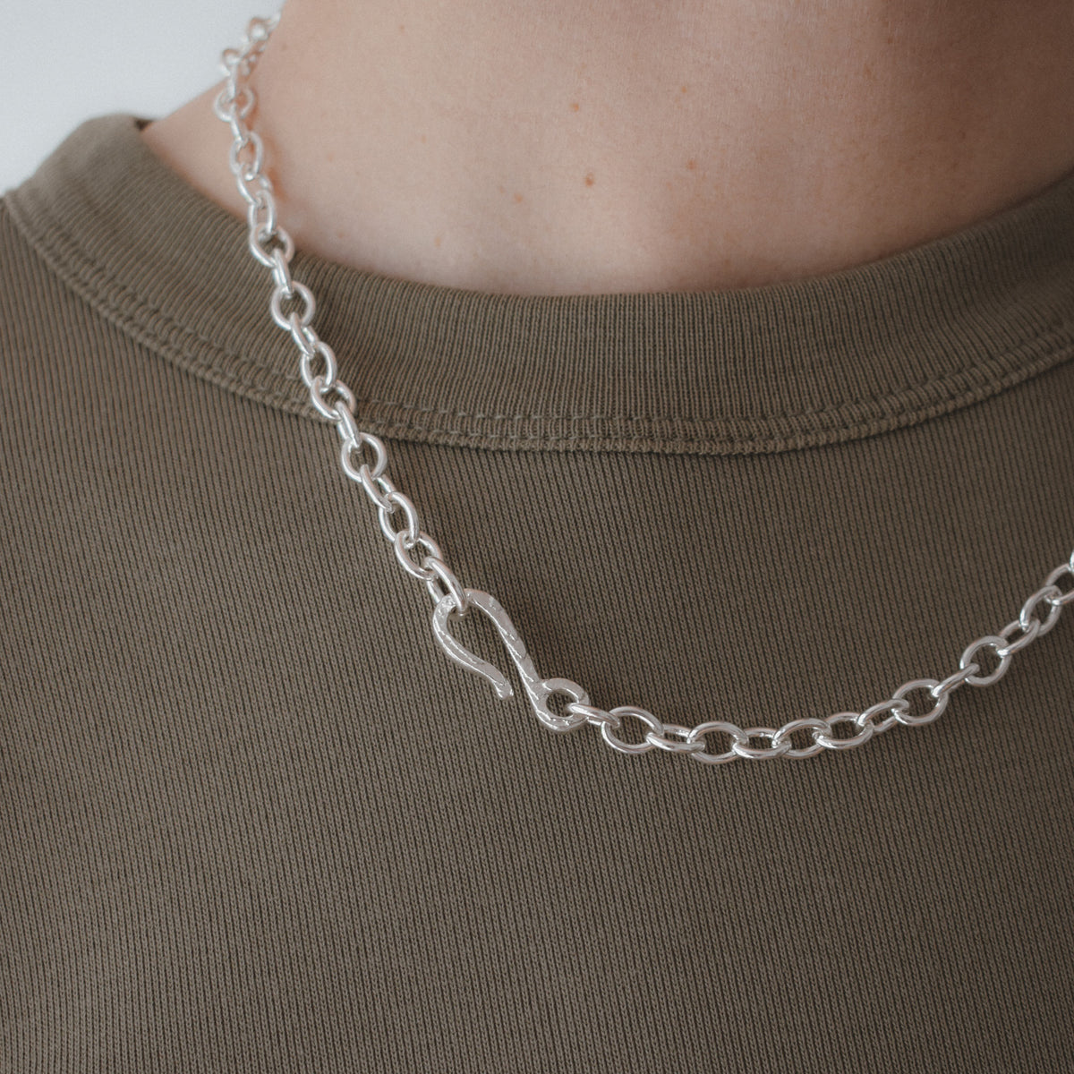 A close-up of a silver chain necklace with a polished infinity symbol pendant, worn on the neck of a person wearing an olive green t-shirt.