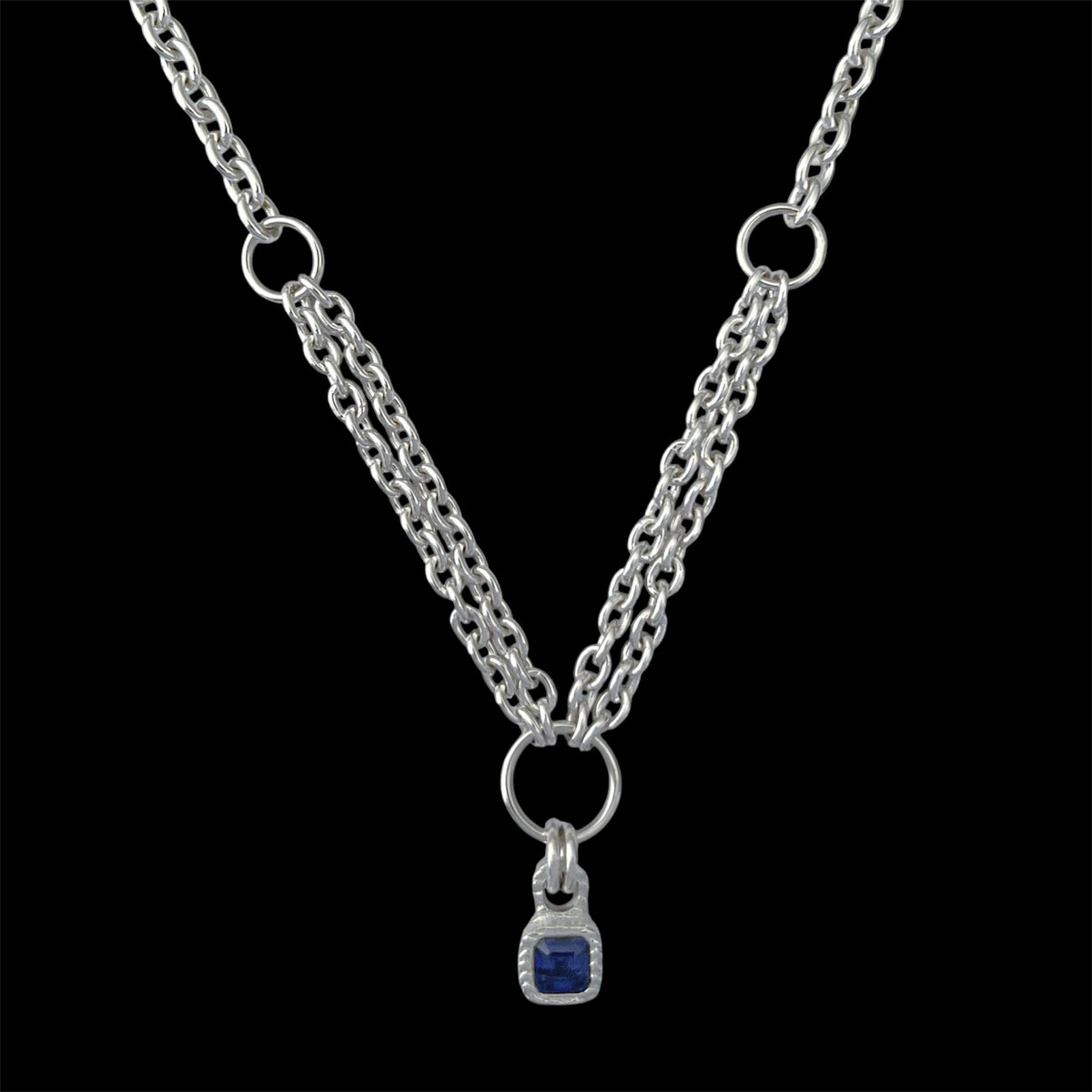A silver-tone double chain necklace with a small square blue gemstone pendant, displayed against a solid black background.