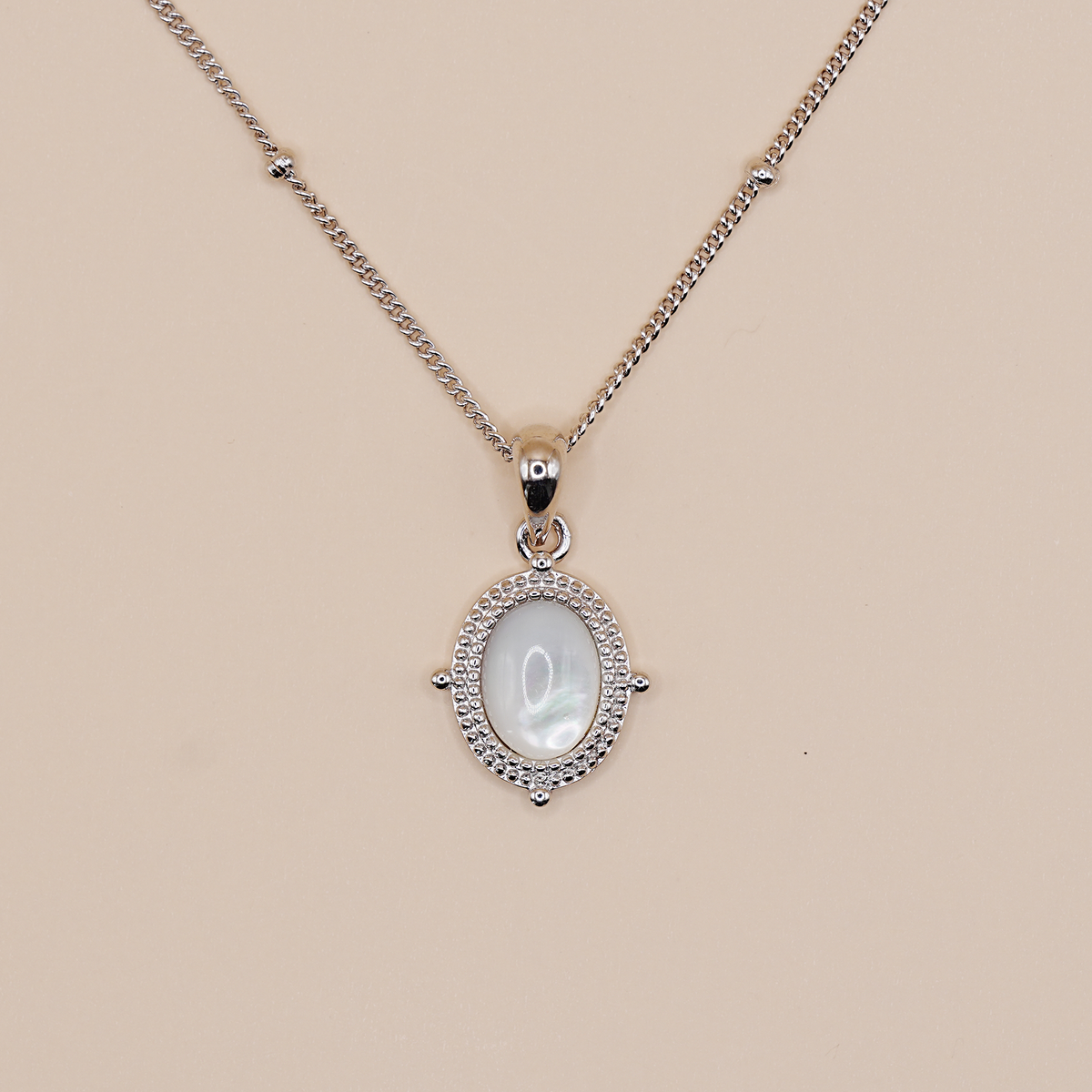 A silver chain necklace with an oval pendant featuring a white mother-of-pearl center stone surrounded by a halo of small clear stones, displayed on a solid beige background.