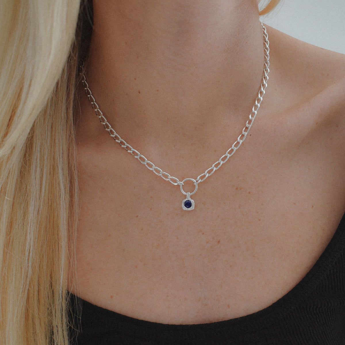 A close-up of a model's neck and chest wearing a silver chain necklace with a small circular ring centerpiece and a tiny dark blue oval gemstone.