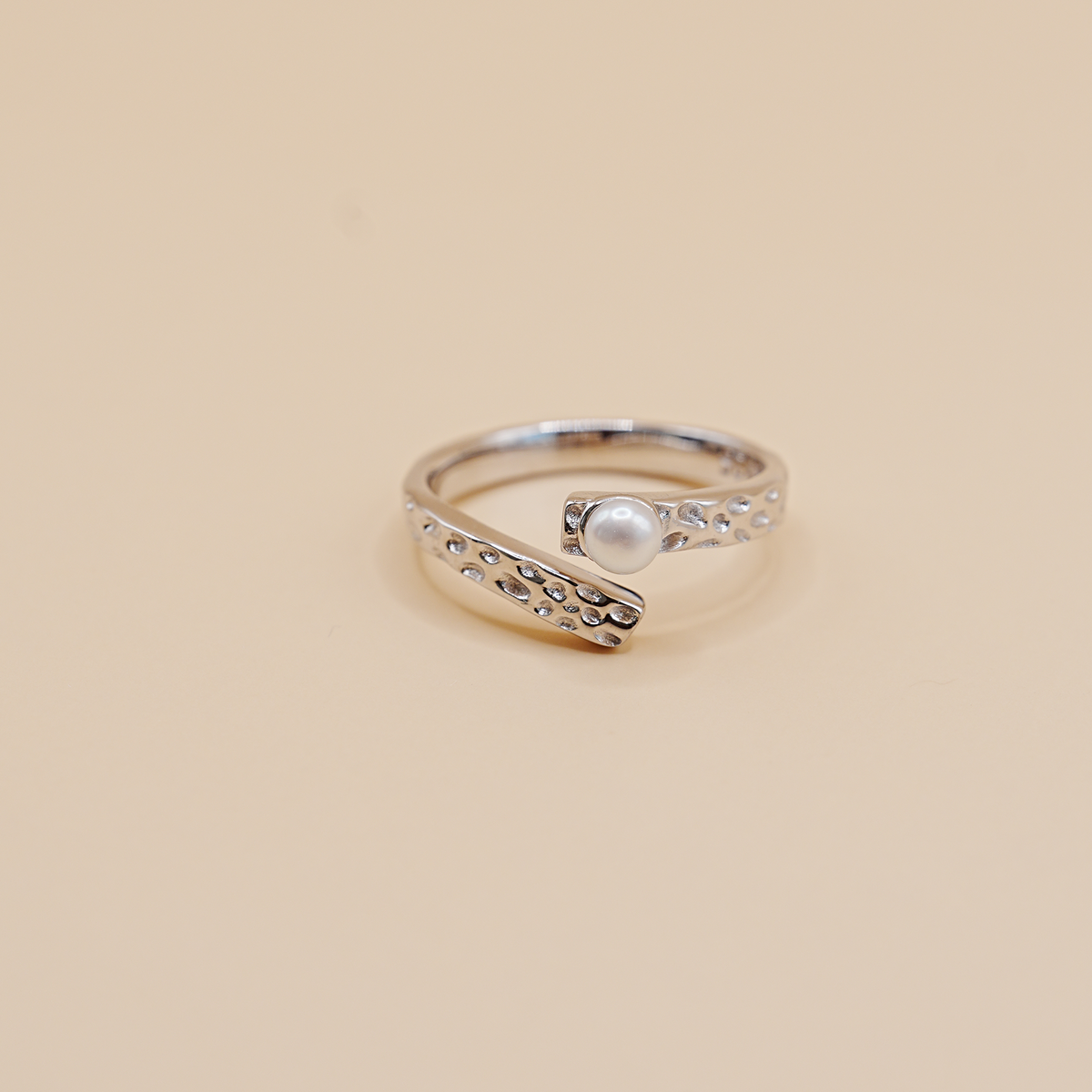 A silver open-band ring with a textured, beaded curved design and a central white pearl, set against a solid beige background.