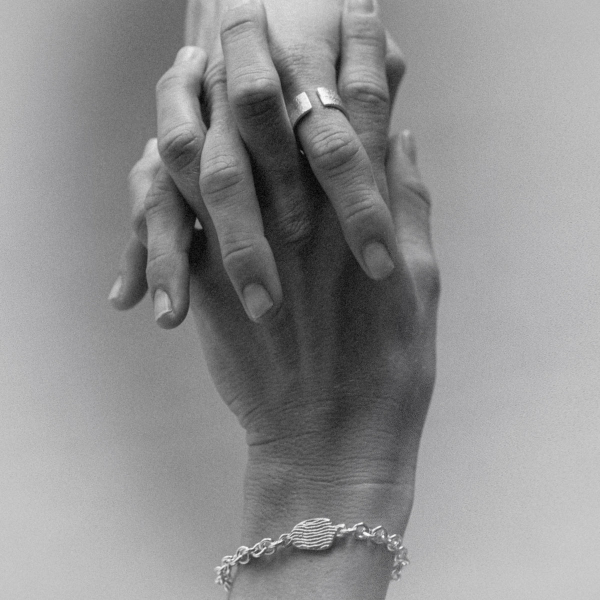 A close-up black and white photograph of two hands clasped together, with a silver ring on one finger and a silver chain bracelet on the wrist.