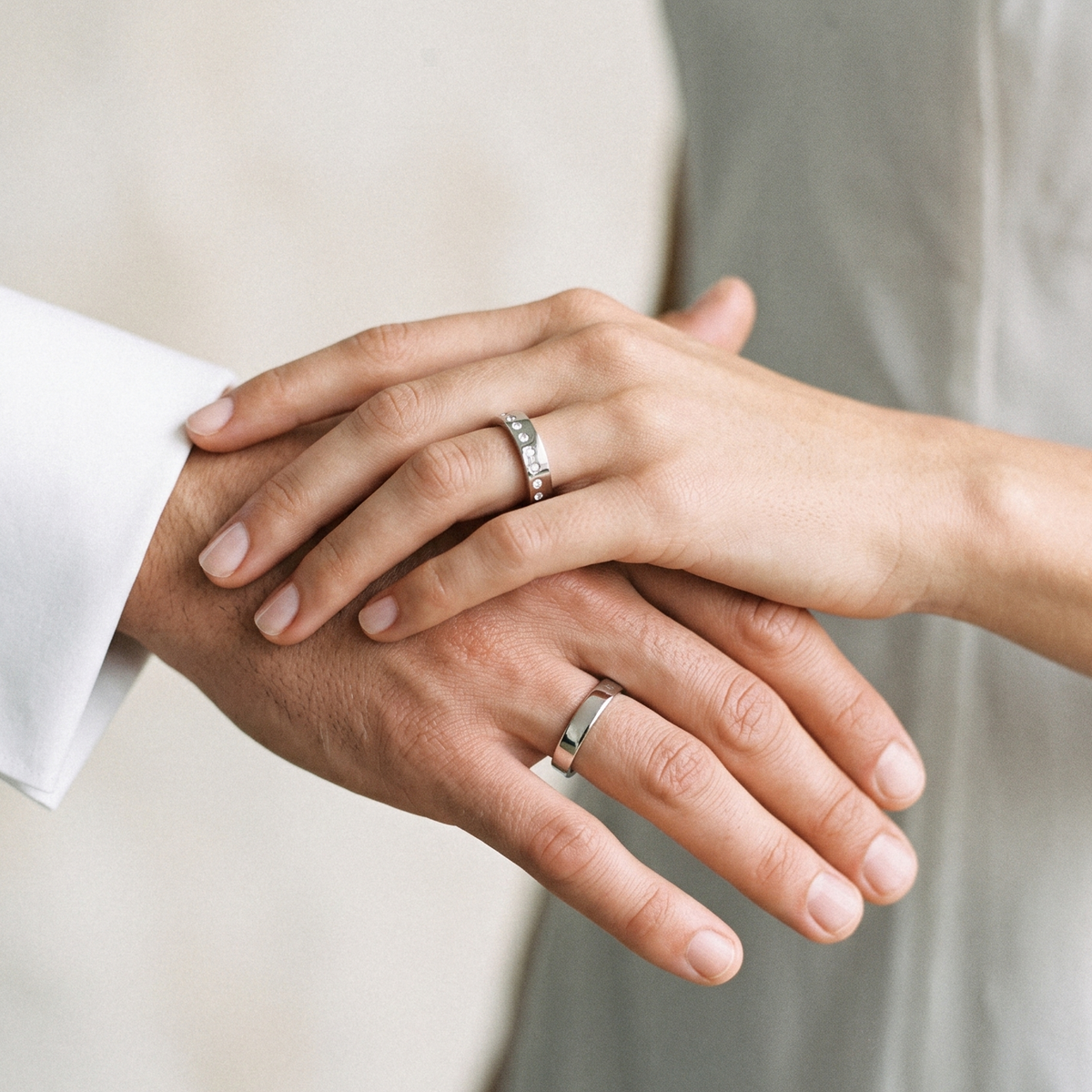 Two hands clasped together, each wearing a simple silver band ring, photographed against a soft neutral background.