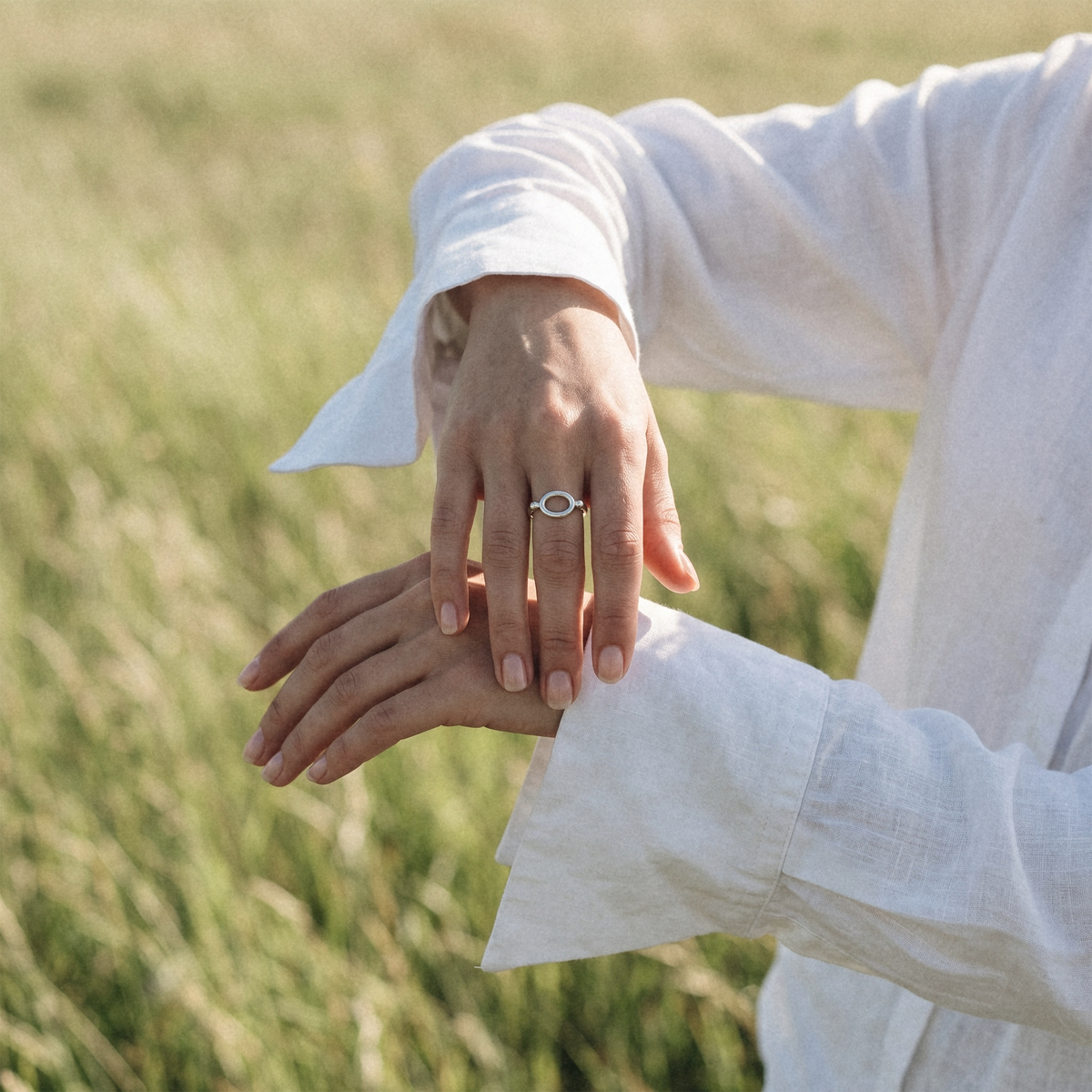 A close-up lifestyle shot of a person wearing a delicate silver ring with a small white stone on their index finger, while adjusting their white linen shirt in an outdoor grassy field.