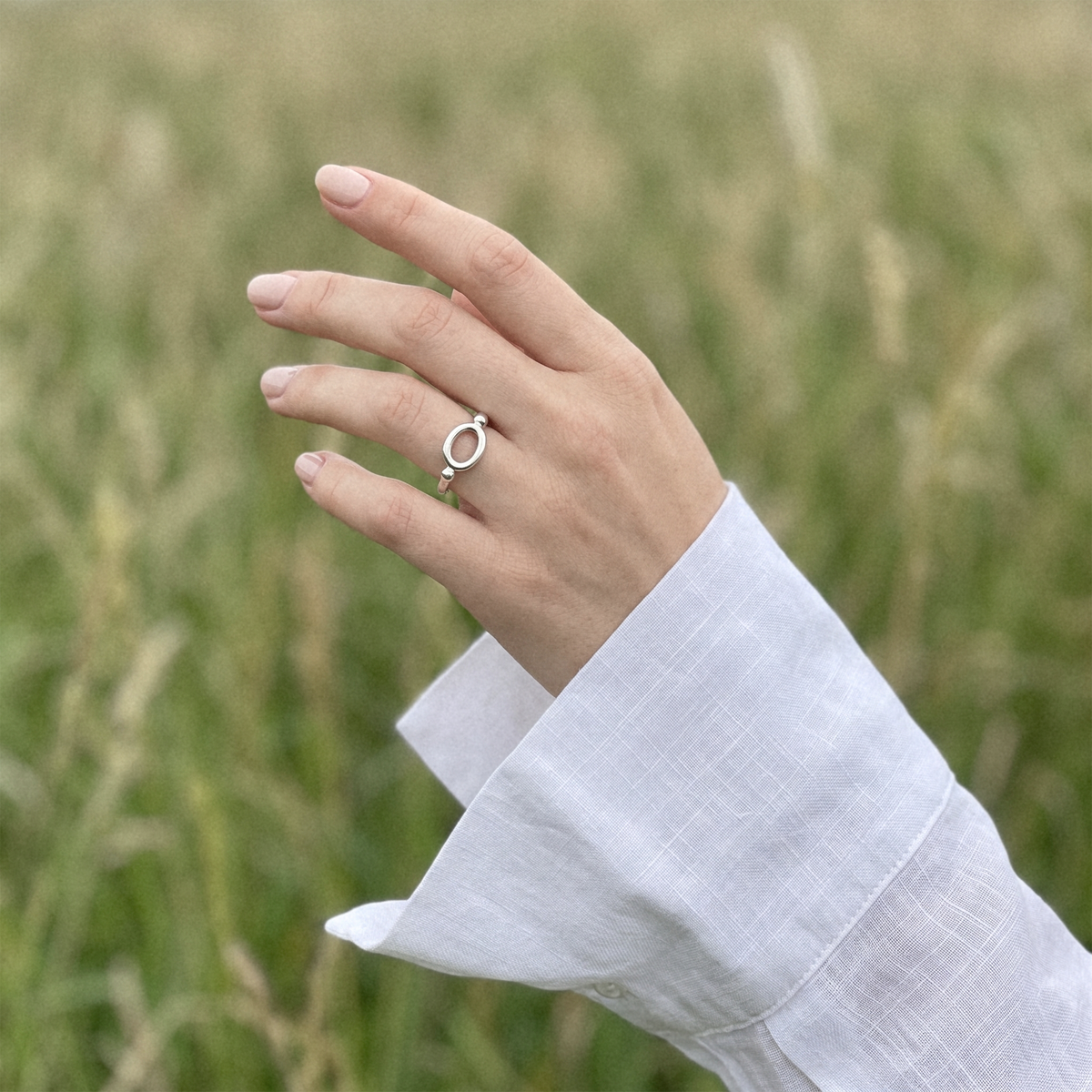 A hand wearing a delicate silver ring with a small circular charm, set against a soft-focus background of a green grassy field. The person is wearing a white linen long-sleeve shirt.