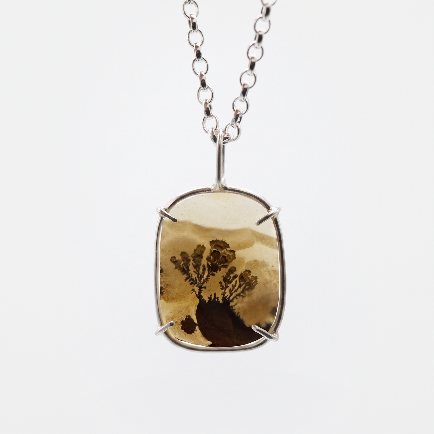 A silver-toned pendant necklace featuring a rectangular bezel with a natural brown and tan tree-in-sand or resin landscape piece, suspended from a silver chain against a light gray background.