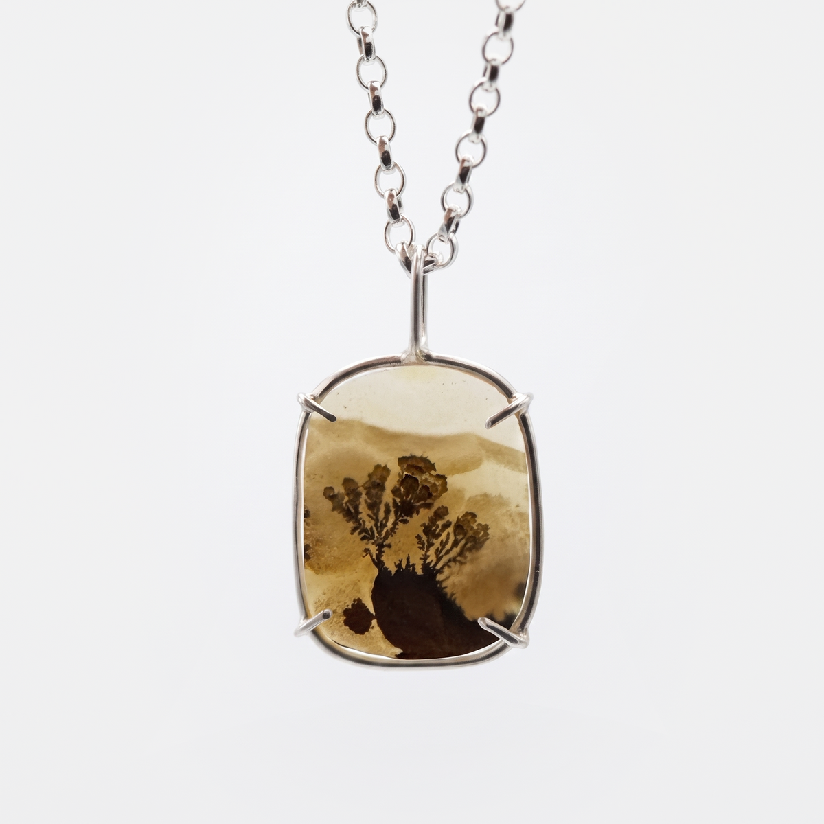 A silver-toned pendant necklace featuring a rectangular bezel with a natural brown and tan tree-in-sand or resin landscape piece, suspended from a silver chain against a light gray background.