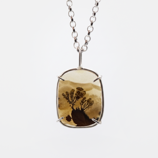 A silver-toned pendant necklace featuring a rectangular bezel with a natural brown and tan tree-in-sand or resin landscape piece, suspended from a silver chain against a light gray background.