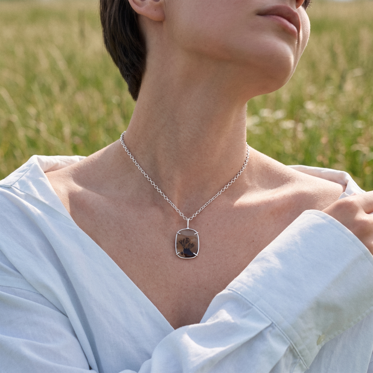 A silver-toned necklace with a rounded rectangular pendant containing a dark brown and black marbled pattern, worn by a model in a light blue shirt against a blurred green field background.