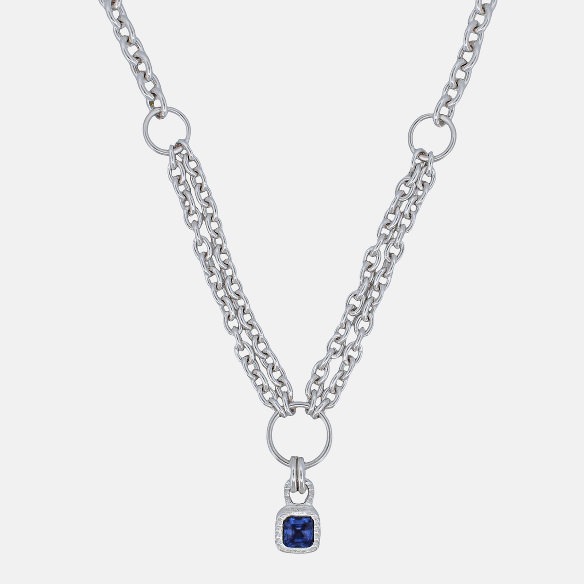 A silver-tone double-strand chain necklace featuring a central ring and a small square pendant with a deep blue gemstone surrounded by a clear crystal border, set against a light gray background.