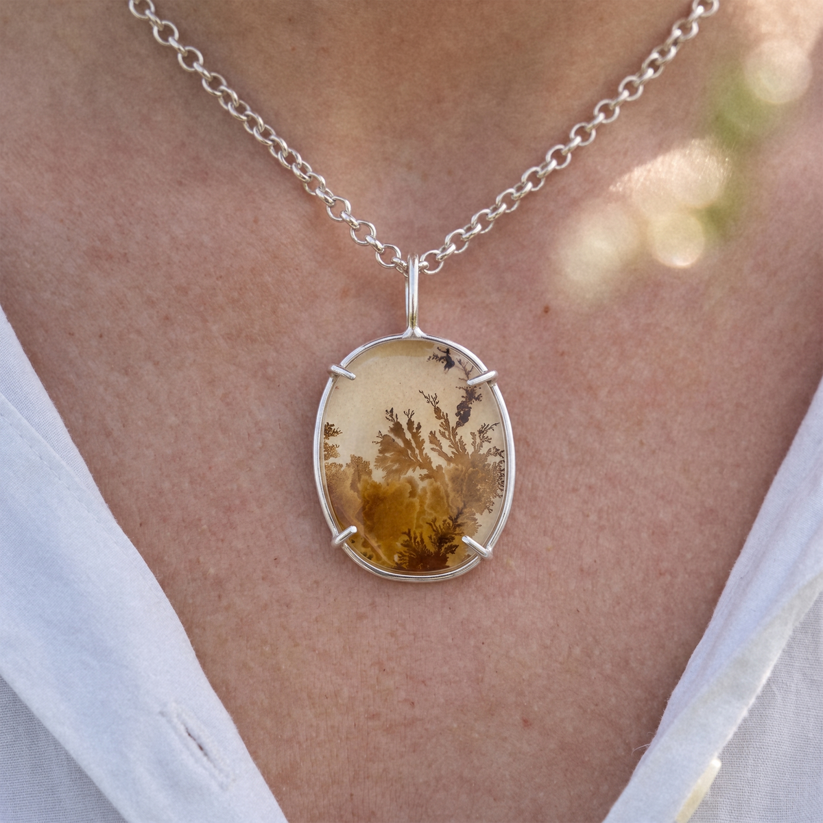 An oval pendant necklace featuring a translucent amber-brown stone with a detailed botanical or coral-like pattern, set in a polished silver-tone bezel and chain.