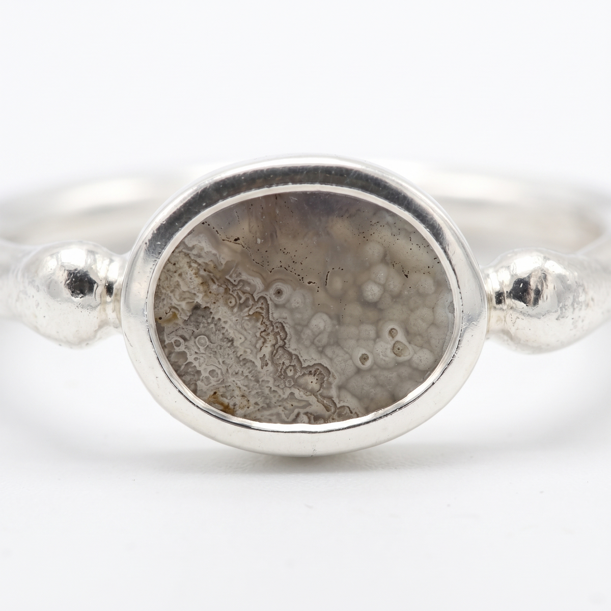 A silver ring featuring an oval cabochon stone with a natural agate or geode pattern in shades of gray, brown, and beige, set on a polished silver band with decorative bead accents on either side.