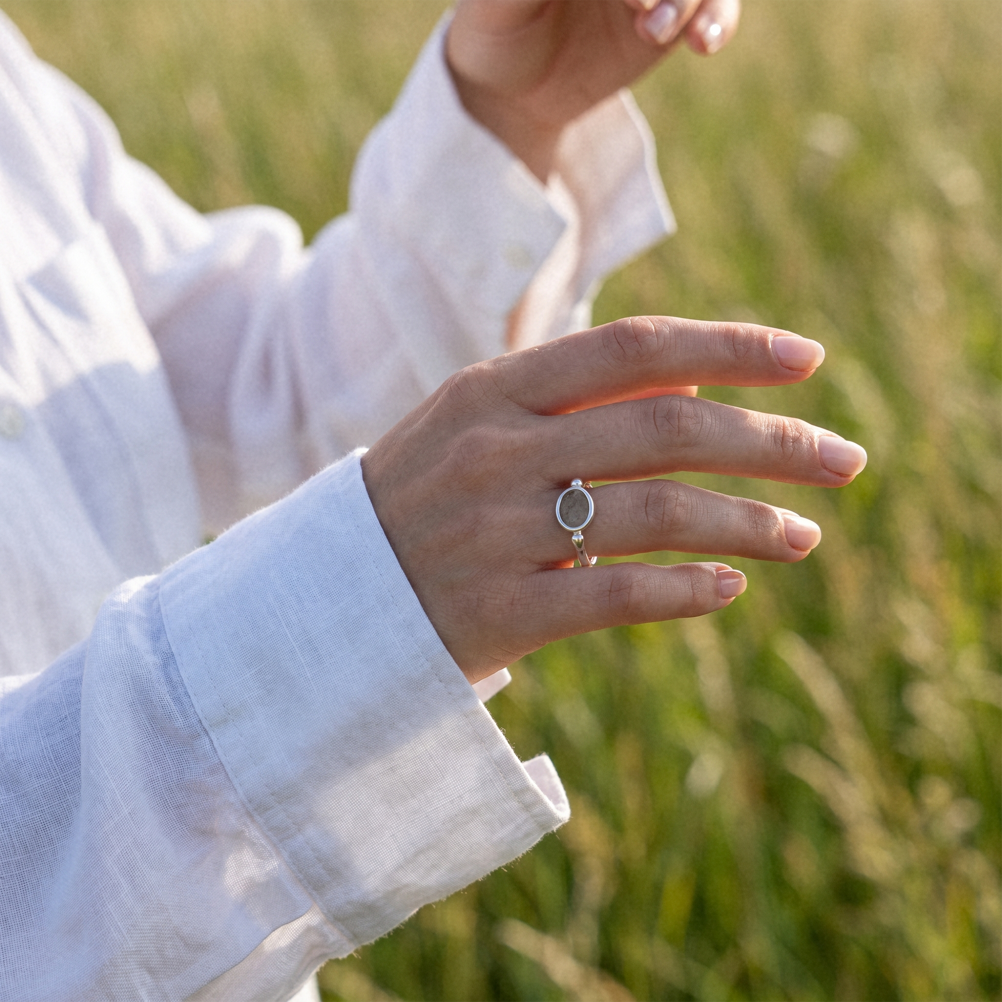 A close-up of a person's hand wearing a silver ring with a circular cutout, set against a soft-focus background of a grassy field.