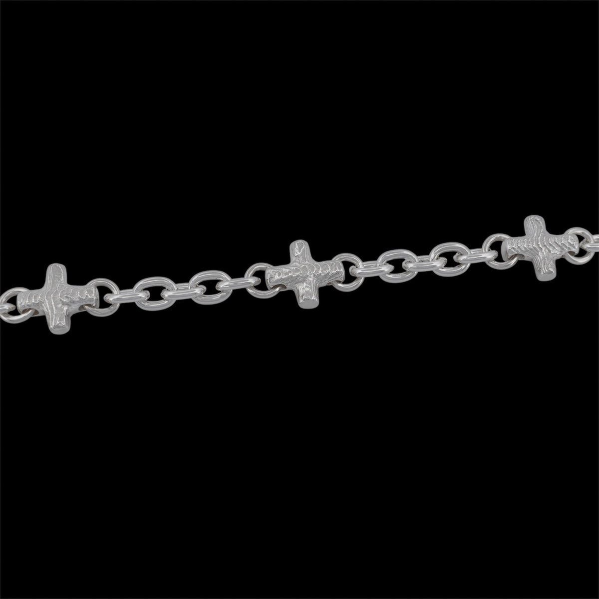 A silver chain bracelet featuring three small, detailed crucifix links spaced evenly along a dark black background.