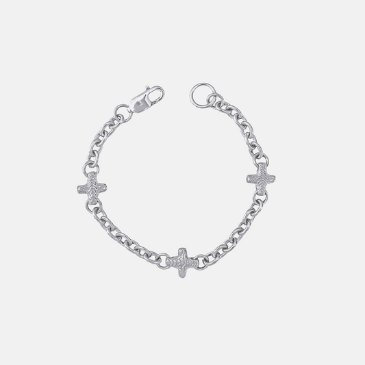 A silver chain bracelet featuring three small cross charms, one of which is adorned with a textured pavé diamond, shown on a light gray background.