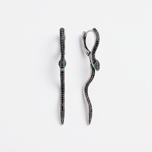 A pair of silver snake drop earrings paved with small black stones, featuring a small green stone accent at the head, shown on a light gray background.