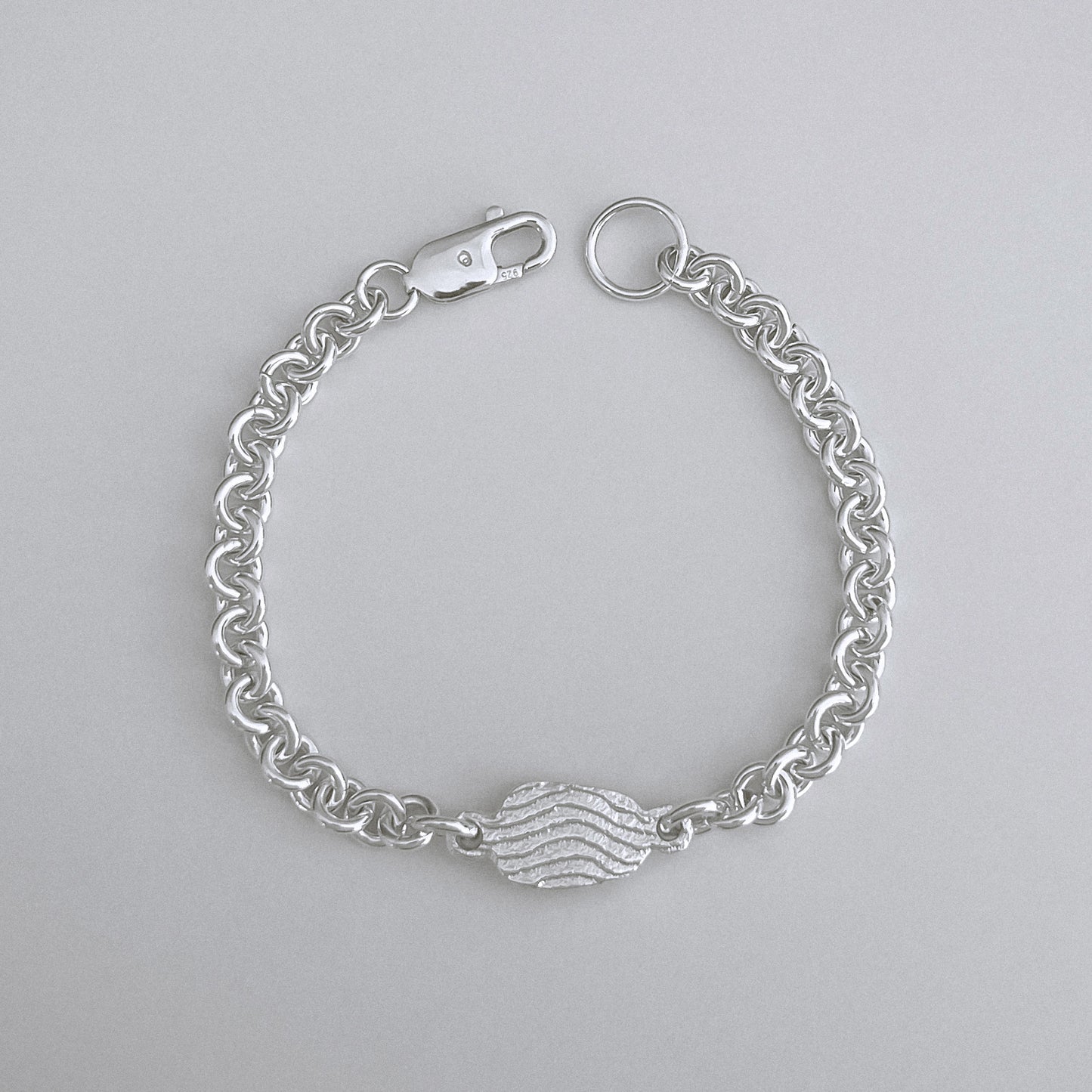 A silver chain bracelet with a central textured heart charm, displayed on a neutral light gray background.