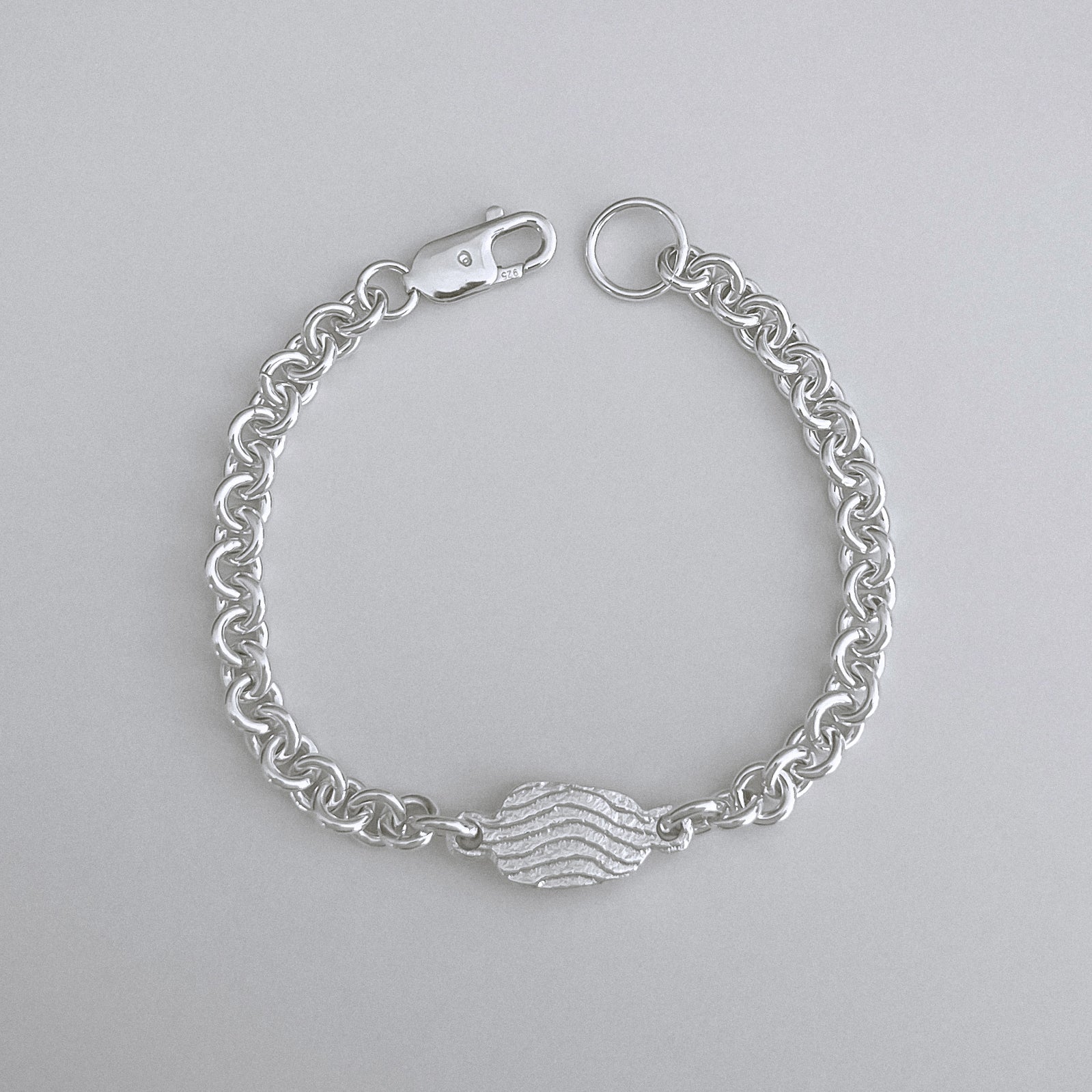 A silver chain bracelet with a central textured heart charm, displayed on a neutral light gray background.