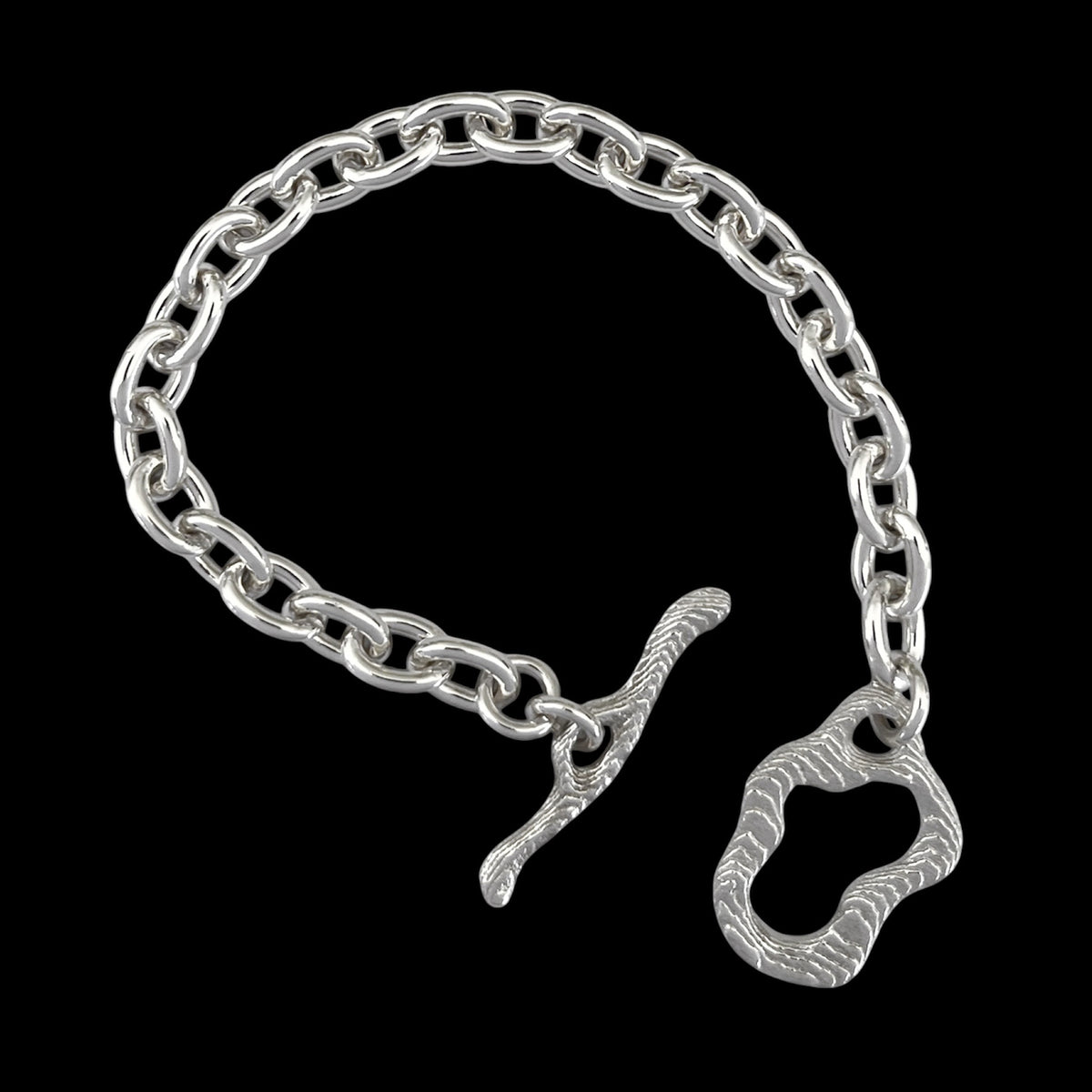 A silver chain bracelet with a unique textured toggle clasp and a matching decorative ring charm, displayed on a solid black background.