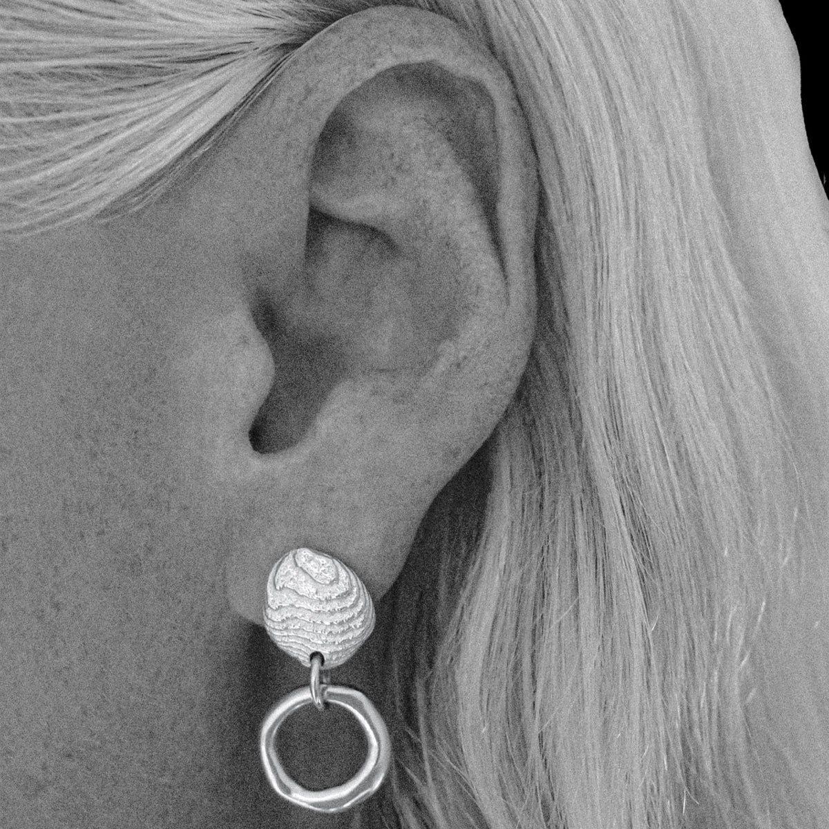 A close-up black and white photograph of a person's ear wearing a textured silver drop earring with a circular dangling element.