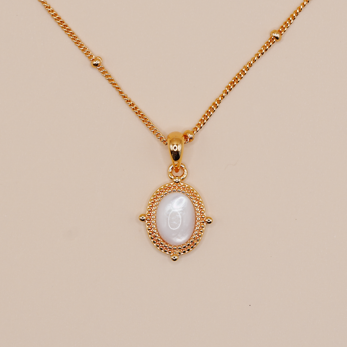 A gold chain necklace with an oval pendant featuring a white mother-of-pearl center and a textured gold border, displayed on a solid beige background.