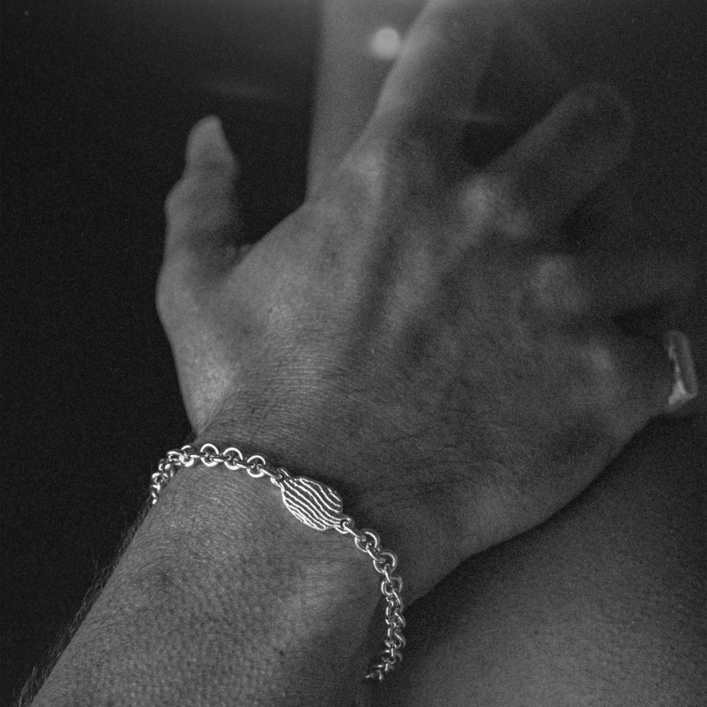 A close-up black and white photograph of a person's hand resting on their leg, wearing a silver chain bracelet with a textured oval centerpiece.