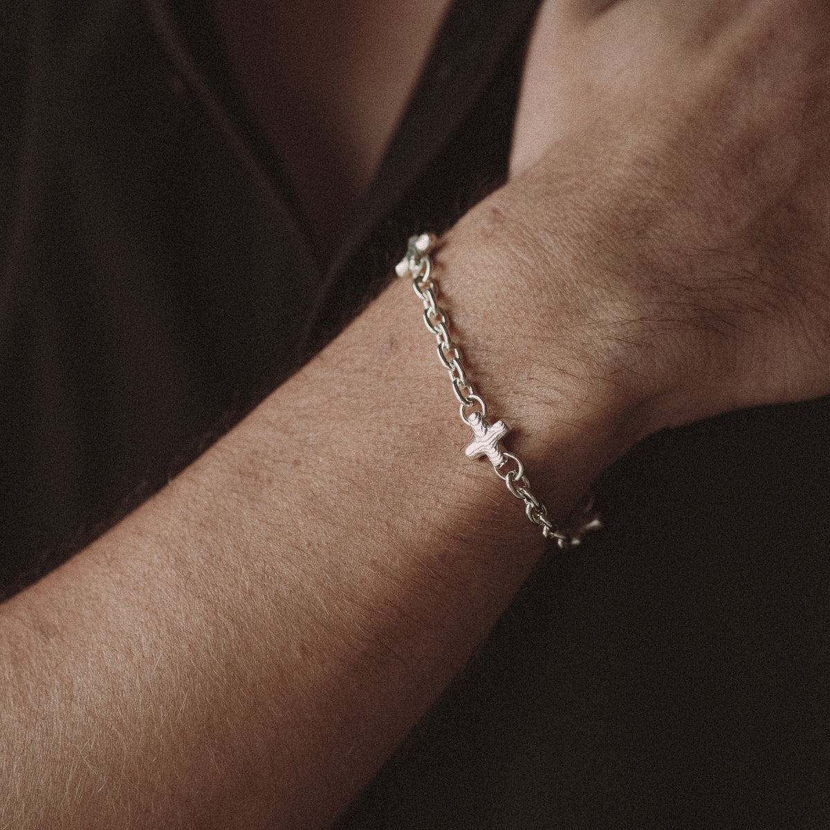A delicate silver chain bracelet with a small cross charm, worn on a person's wrist against a dark brown background.