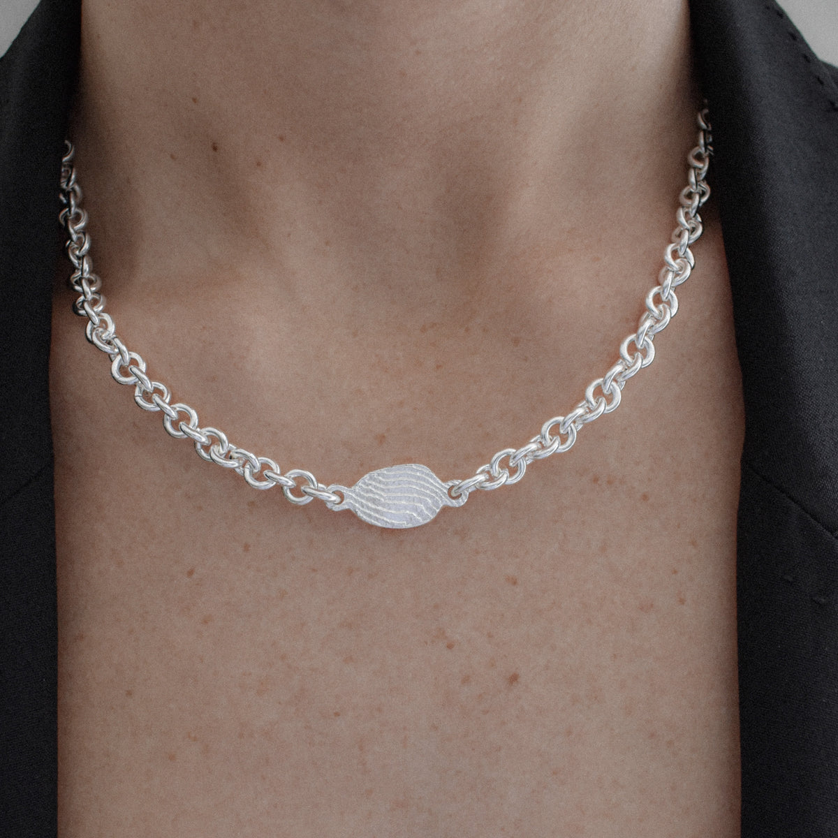 A close-up of a person's neck and chest wearing a silver chain necklace with a textured oval center charm, styled with a black blazer.