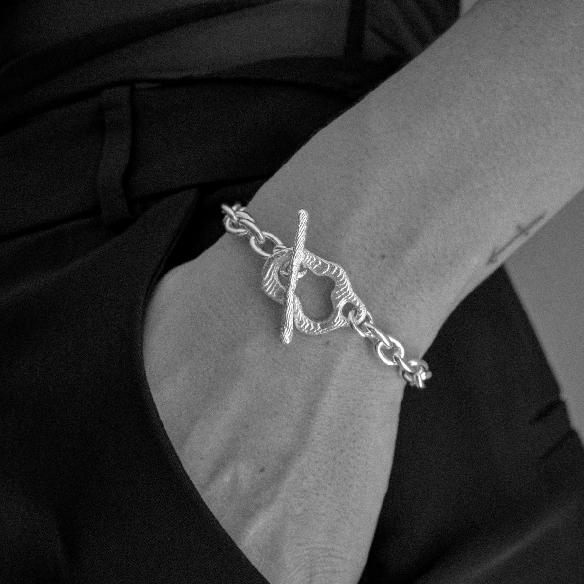 A silver-toned toggle clasp chain bracelet worn on a person's wrist, shown in a black-and-white close-up against dark clothing.