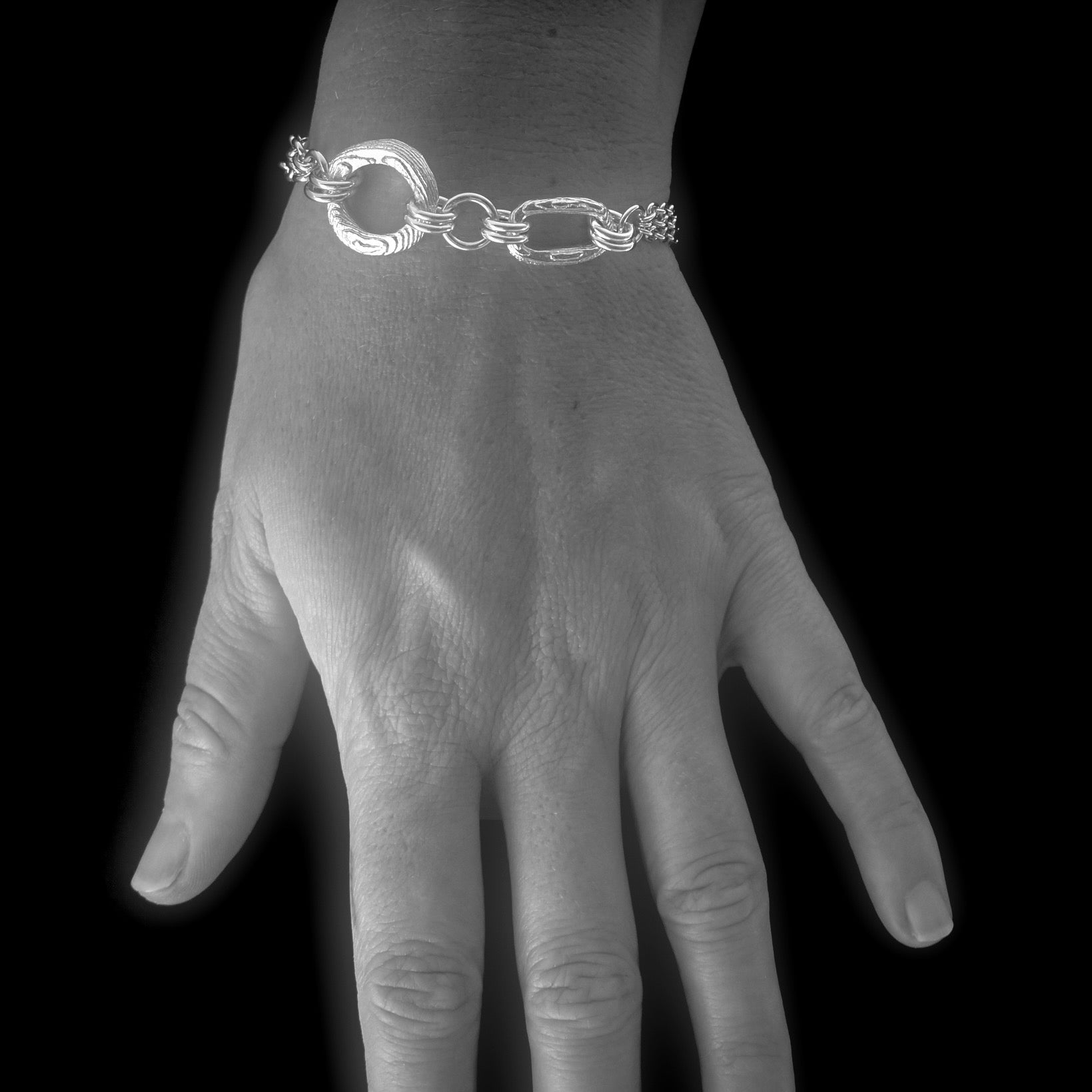 A silver chain bracelet with a textured circular link and elongated oval links, shown worn on a hand against a black background.