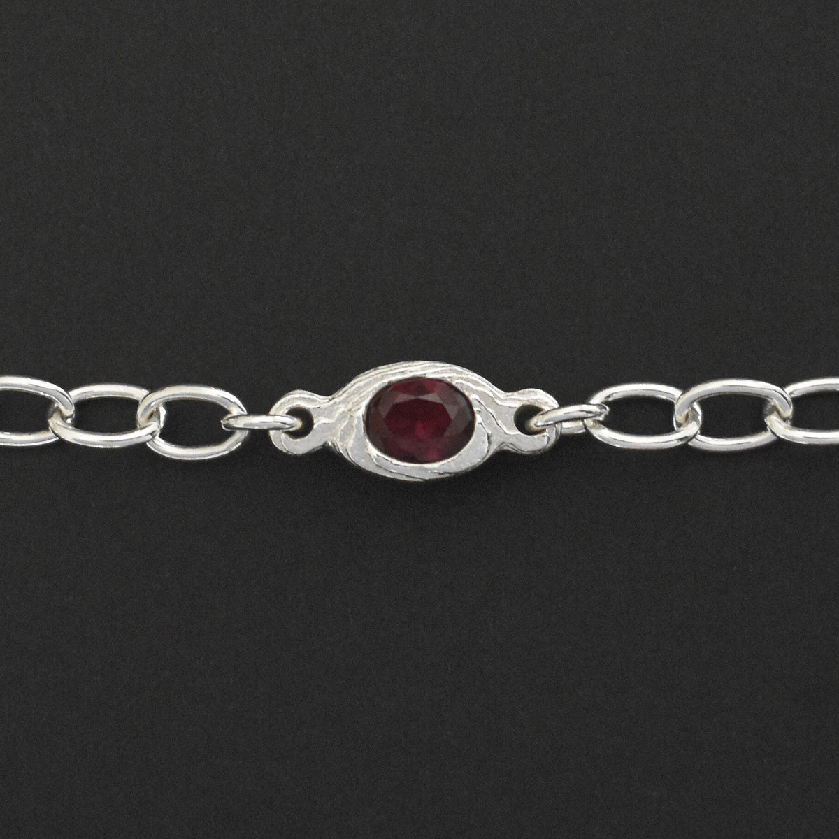 A sterling silver chain bracelet featuring a central oval bezel-set dark red gemstone, shown against a solid black background.
