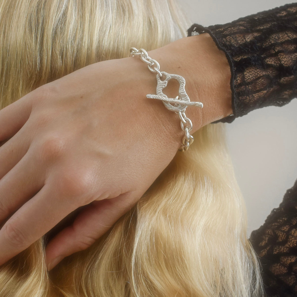 A silver chain bracelet with a toggle clasp worn on a woman's wrist, shown against her blonde hair and a black lace sleeve.