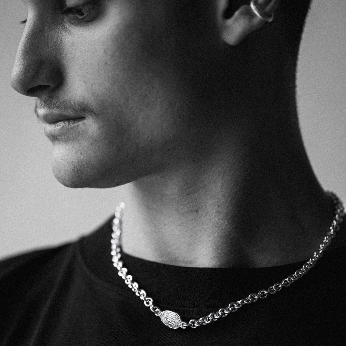 A close-up black and white photo of a man's profile wearing a silver chain necklace with a textured oval pendant.