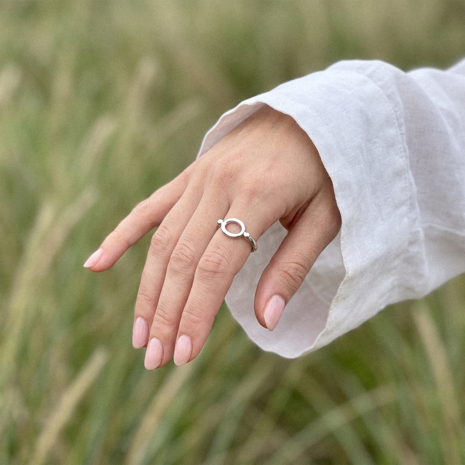 A silver ring with a circular open design worn on a person's ring finger, shown against a soft-focus background of green grass and a white sleeve.