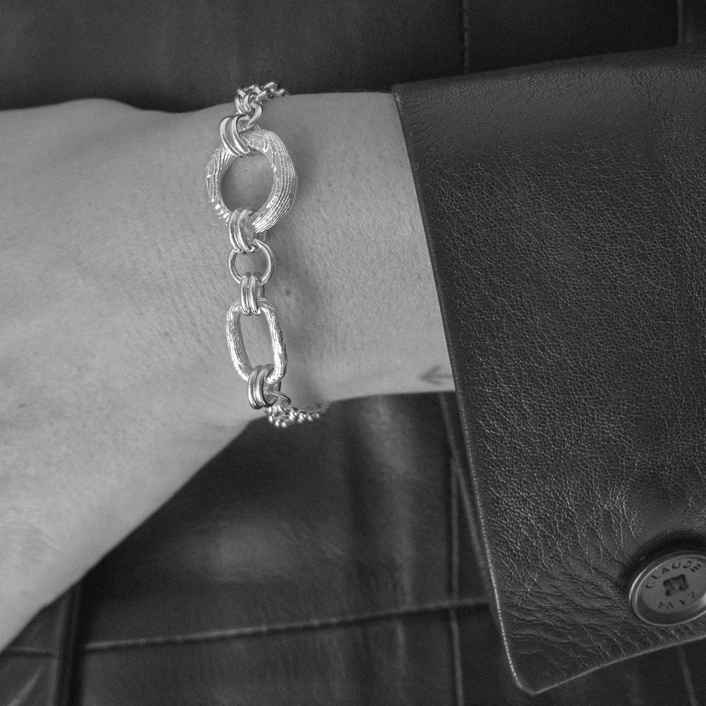 A silver-toned textured link bracelet with oval and elongated links, shown worn on a person's wrist next to a black leather jacket and a black leather accessory.