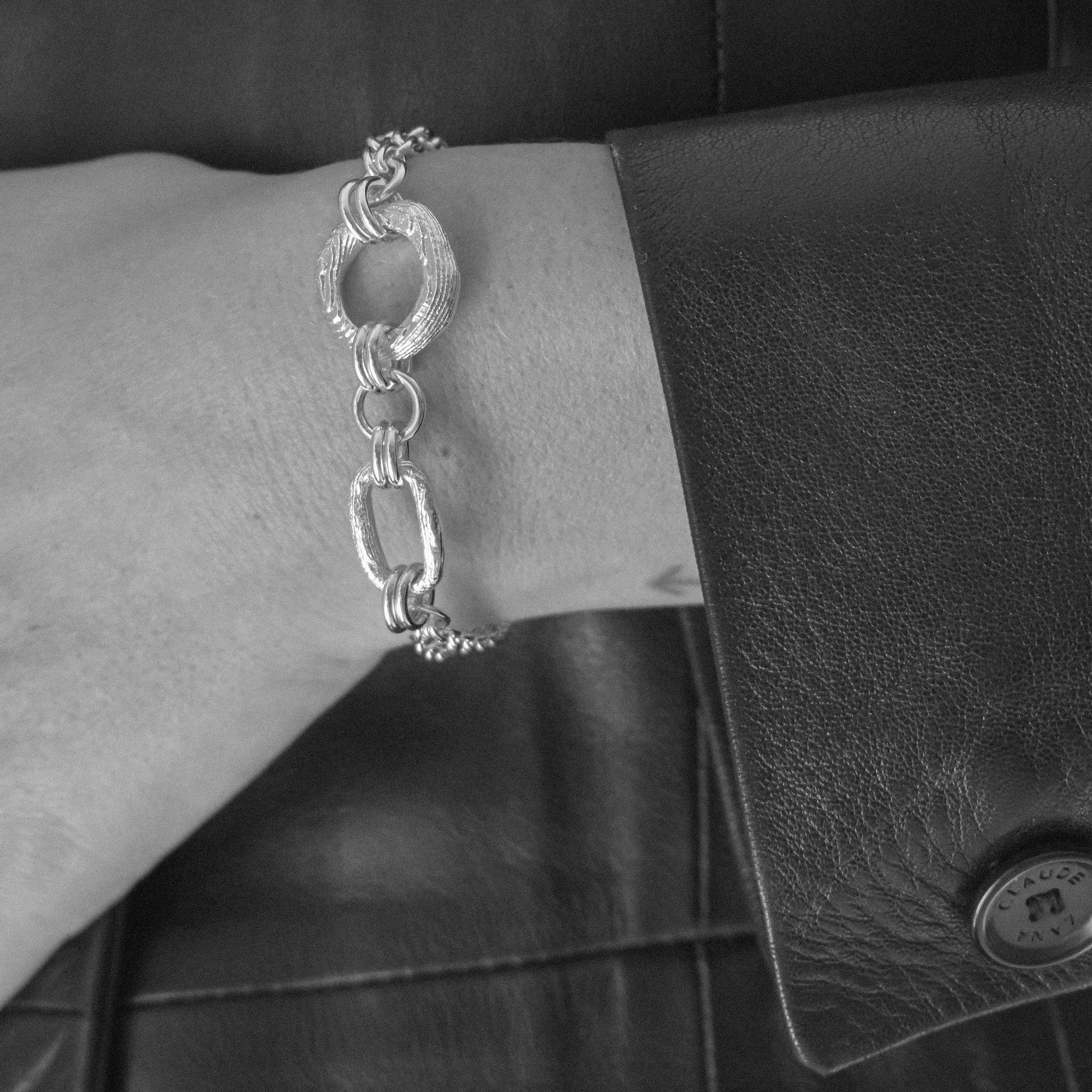 A silver-toned textured link bracelet with oval and elongated links, shown worn on a person's wrist next to a black leather jacket and a black leather accessory.