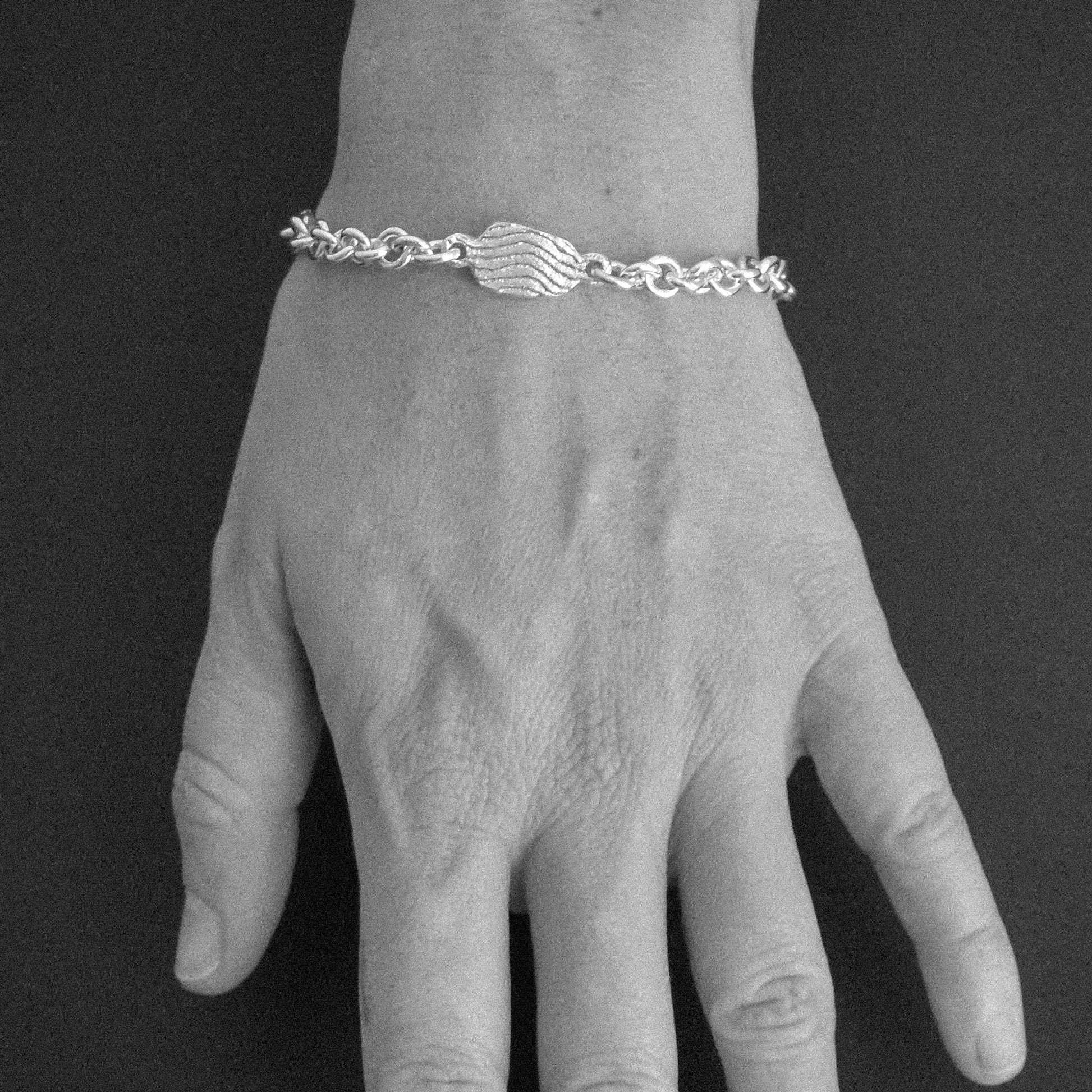 A silver chain bracelet with a central wave-like charm, shown worn on a hand against a dark gray background.