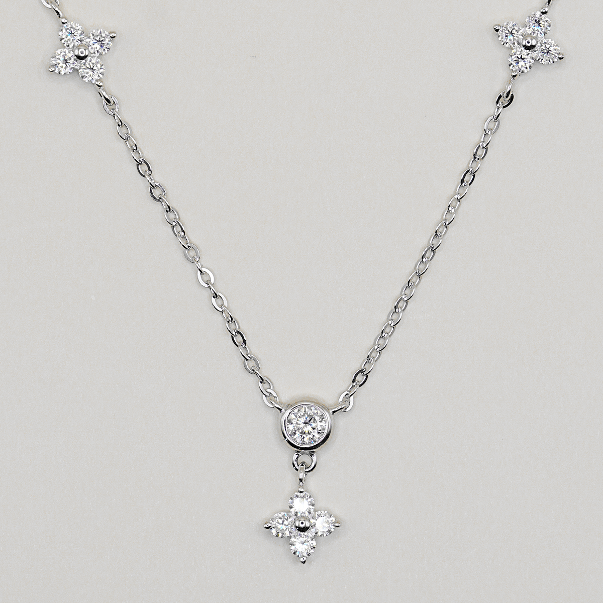 A silver-tone chain necklace featuring a central round bezel-set clear stone and a dangling four-petal flower charm, with matching flower-shaped end pieces on the chain, shown on a light gray background.