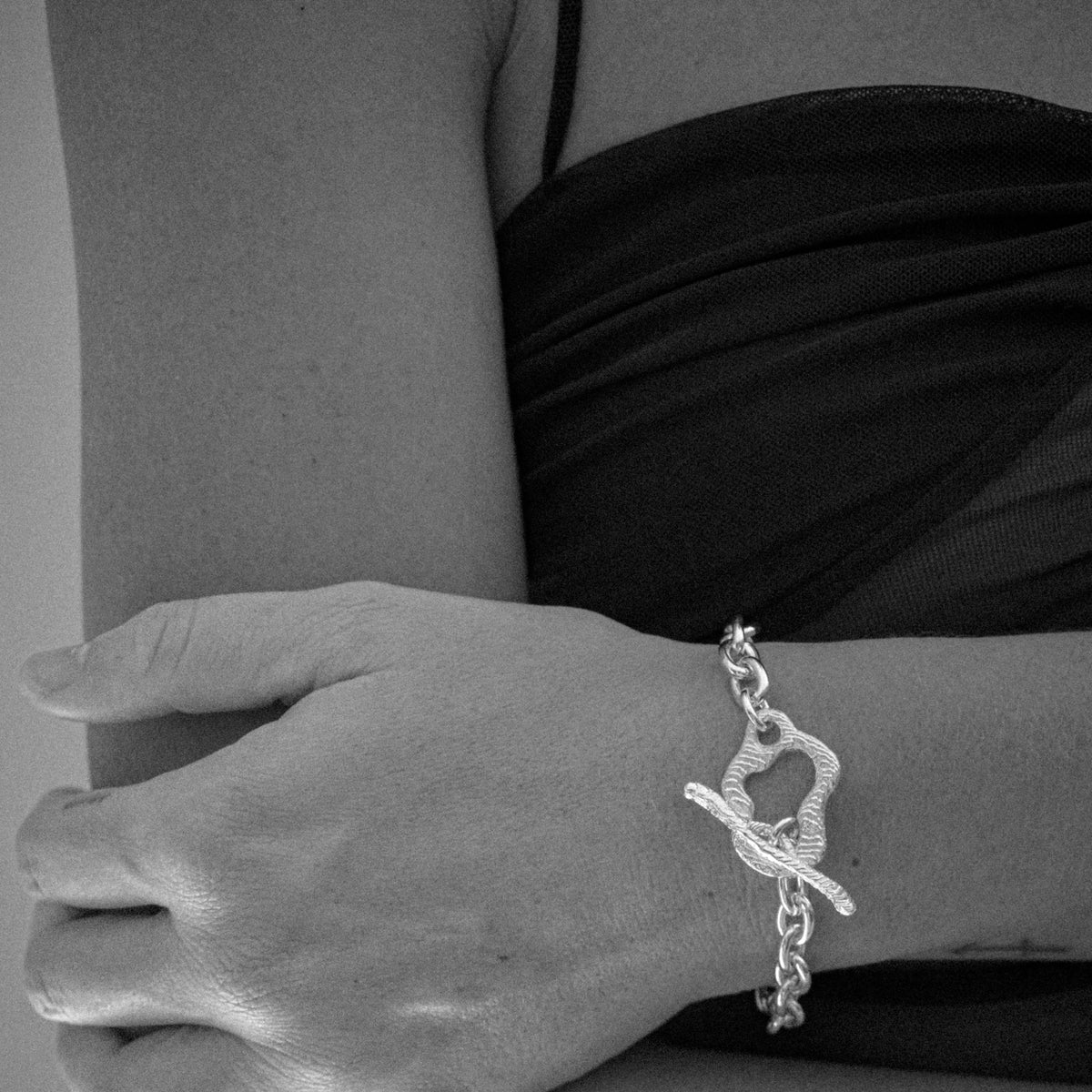 A close-up of a person's wrist wearing a silver chain bracelet with a toggle clasp, shown in black and white.