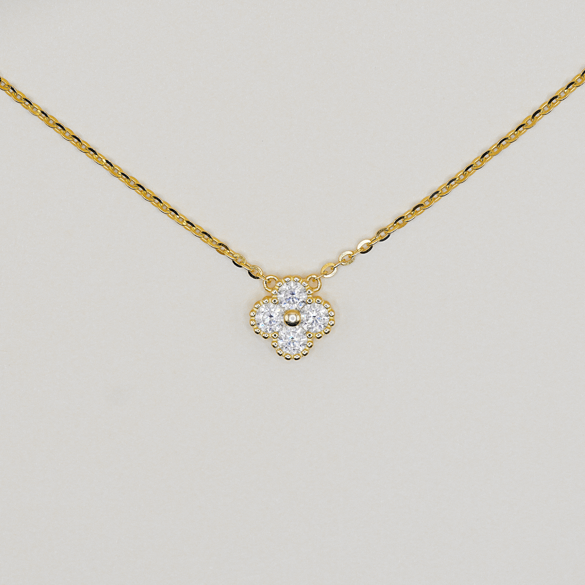 A delicate yellow gold chain necklace with a small four-petal flower pendant. The pendant is set with clear gemstones and a central gold bead, photographed against a neutral light gray background.